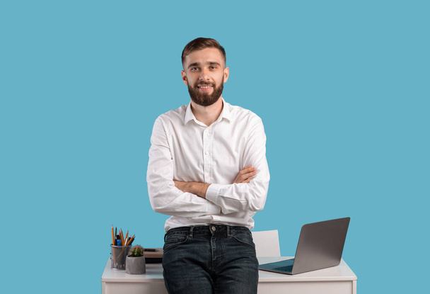 Happy young businessman standing with crossed arms near desk with laptop computer on blue studio background - Фото, изображение