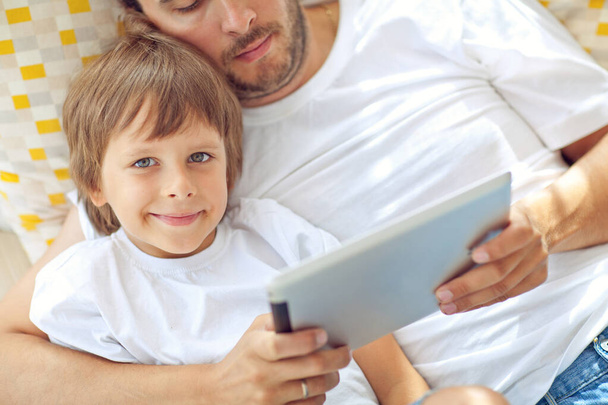 Son and father are playing a computer game on a tablet Closeup of young boy sitting with father and using mobile phone in modern sunny place. - 写真・画像