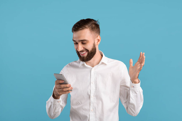 Усміхнений бородатий хлопець використовує смартфон, серфінг в Інтернеті, спілкуючись онлайн на синьому фоні студії
 - Фото, зображення