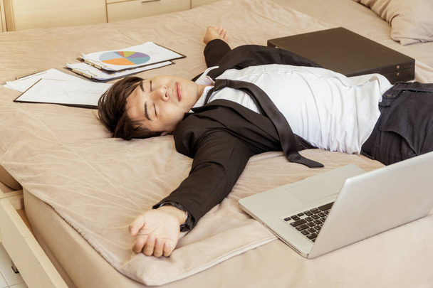Önünde bir dizüstü bilgisayarla yatakta çalışan genç adamın yorgunluğu. Asyalı yakışıklı işadamı yatakta yorgunlukla uyuyor. Önünde defter var.. - Fotoğraf, Görsel