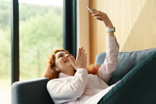 Nuori onnellinen nainen makaa sohvalla kovan työpäivän jälkeen ja tekee videopuhelun - Valokuva, kuva