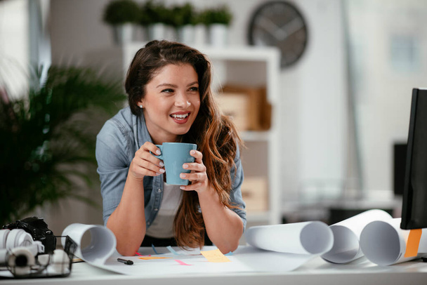 Молода випадкова жінка п'є каву в офісі. Приваблива жінка-менеджер сидить за столом після важкого робочого дня
 - Фото, зображення