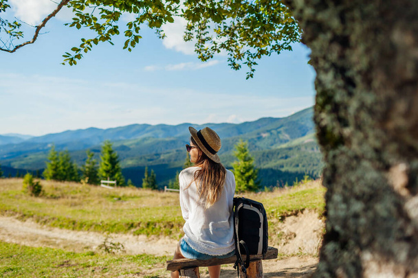 Trip to Carpathian mountains. Woman tourist hiking and relaxing admiring landscape sitting under tree with backpack. Traveling in summer Ukraine - Photo, Image
