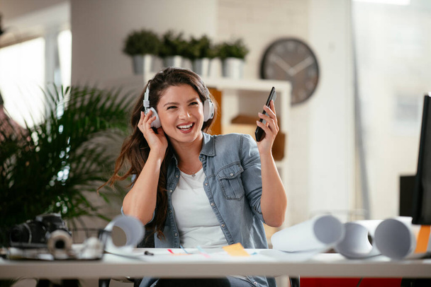 Młoda kobieta w biurze ze słuchawkami. Młoda bizneswoman korzystających w ulubionej piosence przez słuchawki i śpiew podczas pracy na laptopie w biurze. - Zdjęcie, obraz