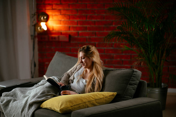 Молодая красивая девушка читает книгу на диване дома. Женщина читает книгу в уютной квартире. - Фото, изображение