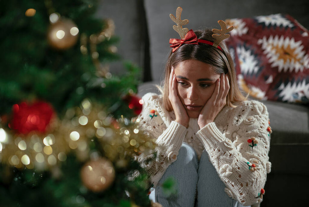 若い悲しい女性はクリスマスツリーの近くに座っている。クリスマス休暇中の憂うつな女性家で. - 写真・画像