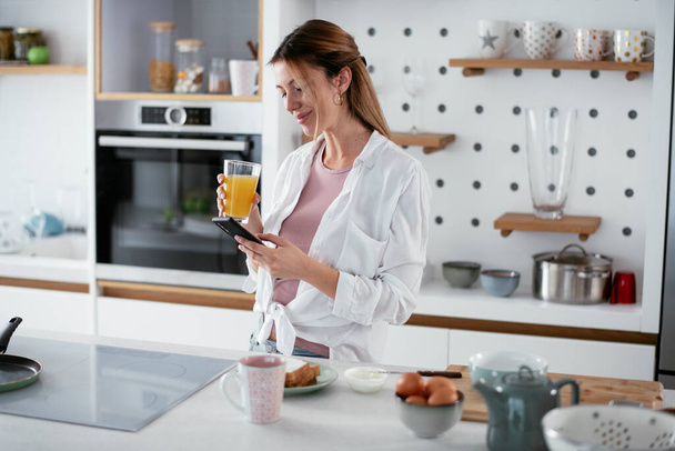 Woman in kitchen. Beautiful woman using phone in kitchen. - Photo, Image