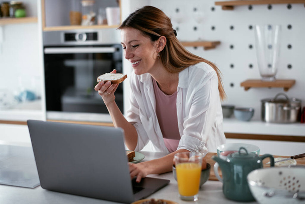 Genç bir kadın laptopla çalışırken kahvaltısının tadını çıkarıyor. - Fotoğraf, Görsel