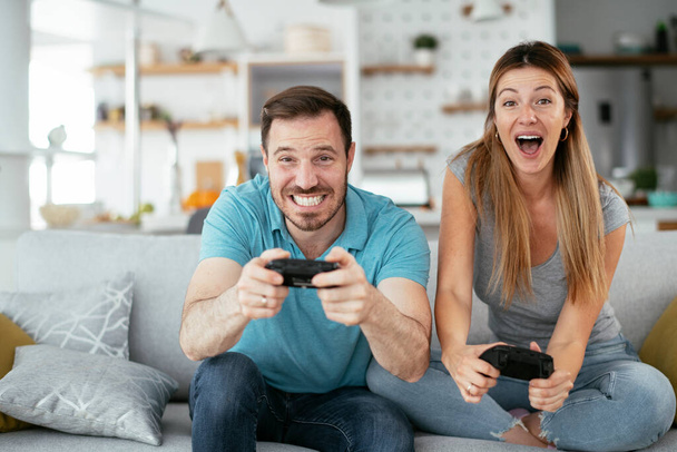 Σύζυγος και σύζυγος παίζουν βιντεοπαιχνίδι με τηλεχειριστήρια στο σαλόνι. Αγαπημένο ζευγάρι παίζουν βιντεοπαιχνίδια στο σπίτι. - Φωτογραφία, εικόνα