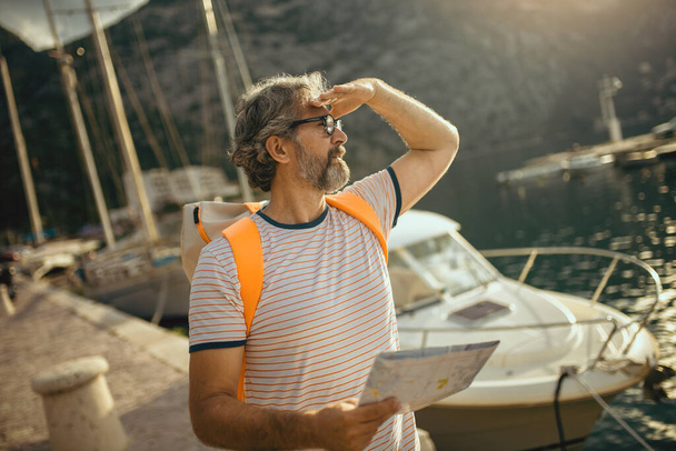Lachende toerist volwassen man staan met kaart en rugzak in de buurt van de zee. - Foto, afbeelding