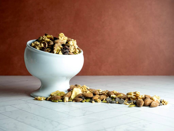 White raised bowl fill of trail mix, nuts and dried fruit spilling out onto the herringbone counter with a terra-cotta wall behind.  Peanuts, granola, pecans, walnuts, banana slices, cranberry. - Foto, Imagen