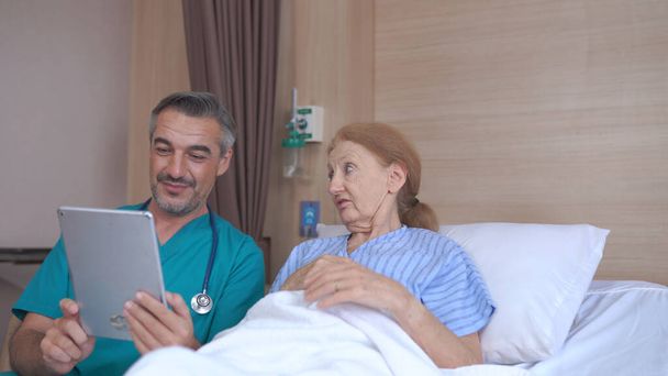 Arzt spricht mit Seniorin mit Tablette im Krankenhausbett - Foto, Bild