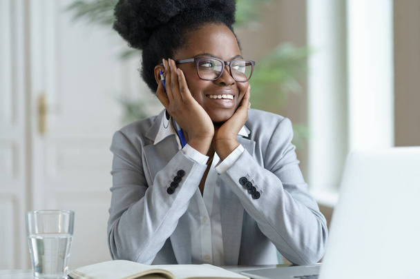Glimlachende Afro vrouw in blazer dragen bril werken op laptop computer thuis kantoor, weg te kijken. - Foto, afbeelding