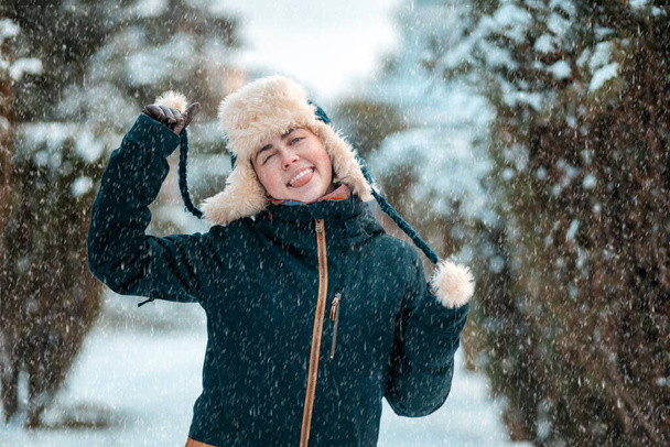 Portret van een jonge vrolijke blanke vrouw met een hoed met oorkleppen en een jasje, die de touwtjes van de hoed in haar handen houdt en vurig haar tong laat zien. Winterseizoen, sneeuwval. - Foto, afbeelding