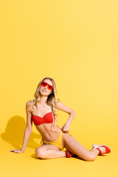 coquettish woman in sunglasses and swimwear looking at camera on yellow - Fotografie, Obrázek