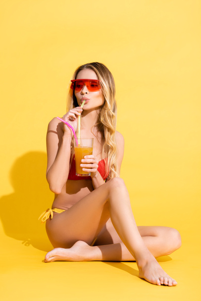 barefoot woman in sunglasses and swimsuit drinking refreshing cocktail while looking away on yellow - Fotó, kép