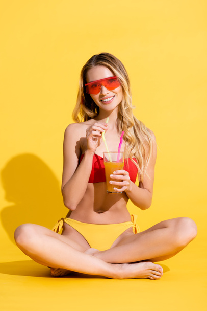 happy woman in swimsuit and sunglasses holding fresh cocktail while sitting with crossed legs on yellow - Fotoğraf, Görsel