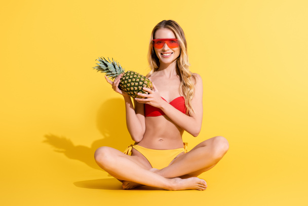 cheerful woman in swimsuit and sunglasses holding whole pineapple while sitting with crossed legs on yellow - Foto, immagini