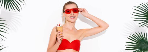 šťastná žena dotýká brýle a usmívá se na kameru, zatímco drží zmrzlinu na bílém, prapor - Fotografie, Obrázek