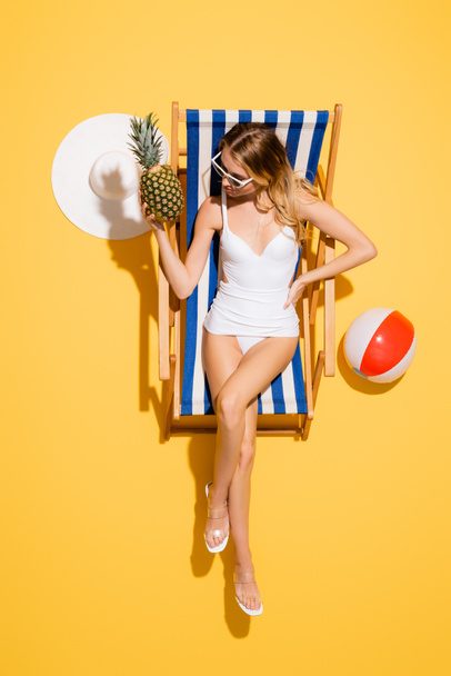 top view of woman with hand on hip holding fresh pineapple while relaxing in deck chair near straw hat and inflatable ball on yellow - Foto, imagen