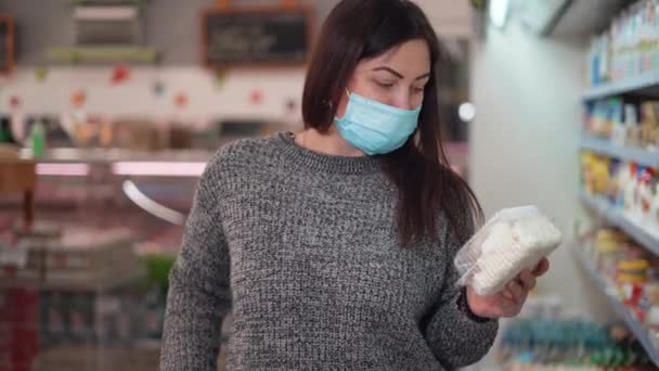 Een vrouw met een beschermend medisch masker koopt voedsel in een supermarkt of supermarkt. De klant legt zuivelproducten, kwark in de mand - Video