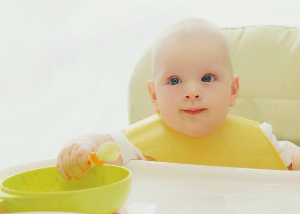 Baby eating with spoon sitting at the table at home - Φωτογραφία, εικόνα