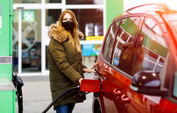 Kobieta nosi maskę medyczną na samoobsługowej stacji benzynowej, trzyma dyszę paliwową, tankuje benzynę podczas pandemii wirusa korony. Ludzie w maskach jako środek zapobiegawczy i ochrona przeciw owadom - Zdjęcie, obraz