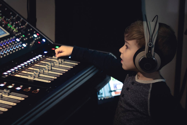 Mały chłopiec ze słuchawkami podkręcającymi głośność podczas edycji muzyki w studiu nagraniowym.  - Zdjęcie, obraz