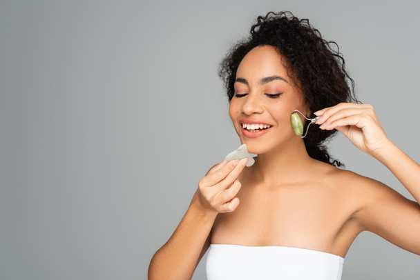 Young african american woman smiling while using jade roller and gua sha isolated on grey  - Φωτογραφία, εικόνα