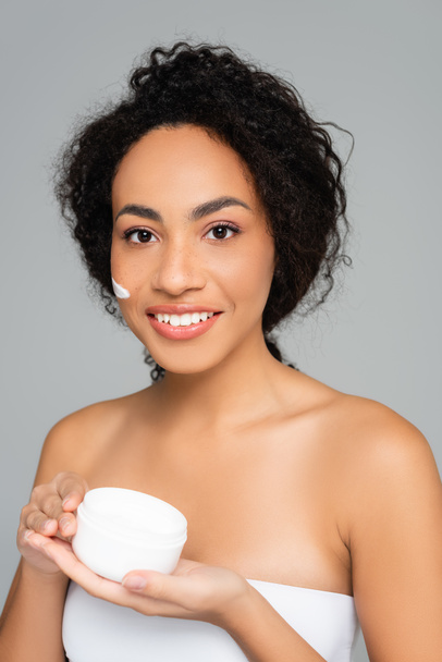African american woman smiling while holding jar with cosmetic cream isolated on grey  - 写真・画像