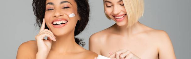 Cheerful multicultural women applying cosmetic cream isolated on grey, banner  - Photo, Image
