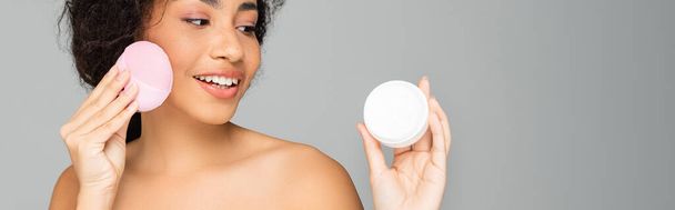 Happy african american woman looking at jar with cosmetic cream while holding silicone cleanser isolated on grey, banner  - Фото, изображение