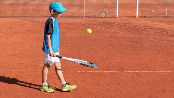 Jogador de tênis infantil joga com bola e raquete na quadra de barro vermelho. Conceito de esportes infantis e atividade física. Crianças divertido jogo desportivo. Fundo, espaço de cópia - Foto, Imagem
