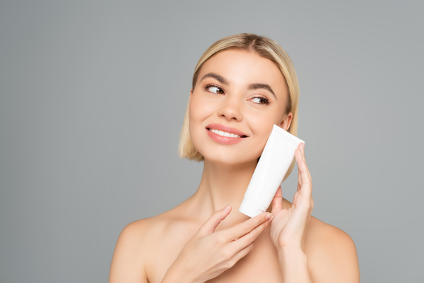 Smiling woman with naked shoulders holding tube with cosmetic cream near face isolated on grey  - Photo, Image