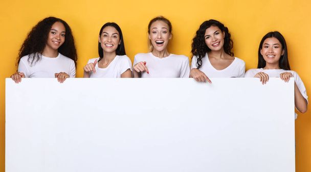 Мультикультурные дамы держат пустую бумажную доску на жёлтом фоне, макет - Фото, изображение