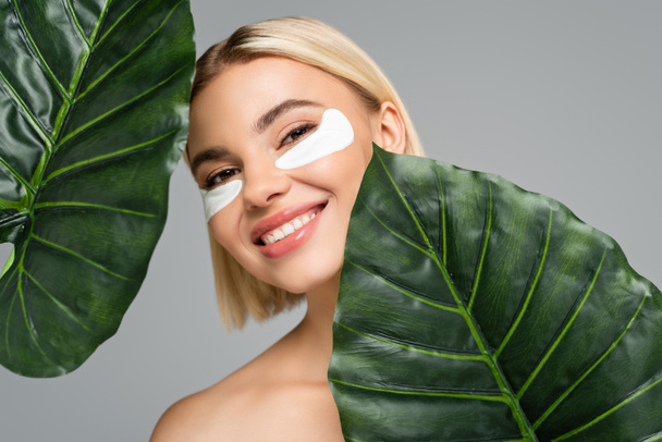 Young woman with eye patches smiling near green leaves isolated on grey  - Foto, imagen