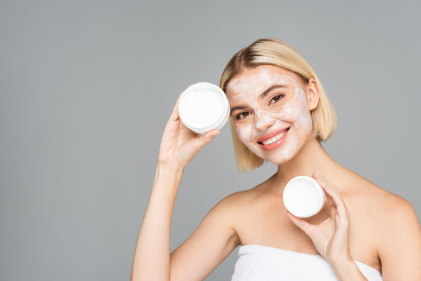 Happy woman in facial mask holding cosmetic cream isolated on grey  - Фото, зображення