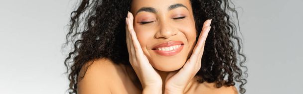 Smiling african american woman with hands near face and closed eyes isolated on grey, banner  - Foto, Bild