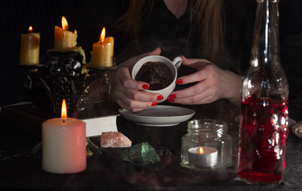 Ennustaminen kahvinporoilla. Ennustajan kädet, kuppi kahvia, kynttilöitä ja kalloja mustalla pöydällä. Velhojen, noituuden ja taikuuden käsite. - Valokuva, kuva
