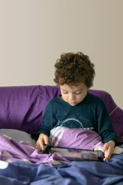 4-5 year old boy with curly hair sitting on the bed with a tablet in his hands playing and entertaining himself in a morning. - Foto, afbeelding