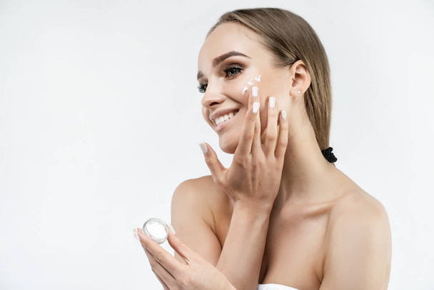 On a white background, a woman who applies cream to her radiant and fresh face. Cosmetology and skin care, healthy skin. Face cream. Place for text. - Photo, Image