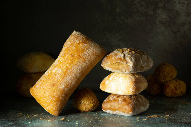 Freshly baked delicious bread, ciabatta, buns on a dark background. Healthy nutrition concept. Balance in food composition. Copy space. - Photo, Image
