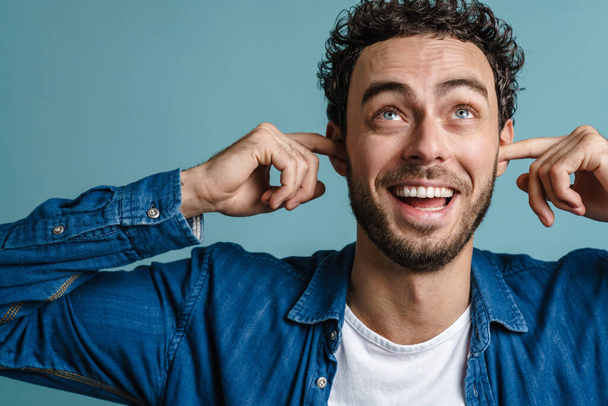 Cheerful handsome guy smiling and plugging his ears isolated over blue background - Foto, afbeelding