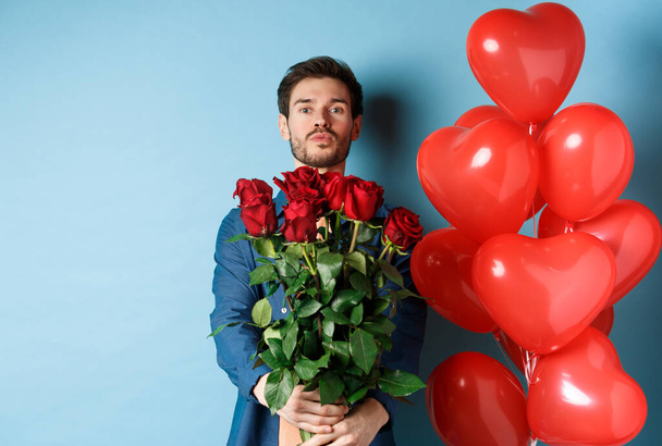 Romantic boyfriend confessing in love and giving bouquet of roses, pucker lips for kiss, standing with heart balloons on blue background - Photo, Image