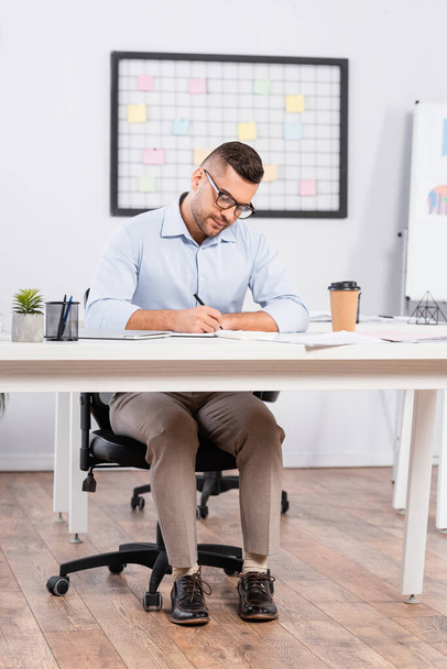 businessman in glasses writing in notebook near paper cup with coffee to go on desk - Foto, imagen