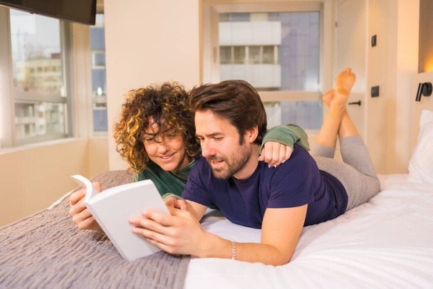 Lifestyle, a young Caucasian couple in pajamas in bed reading a nice book hugging - Фото, изображение
