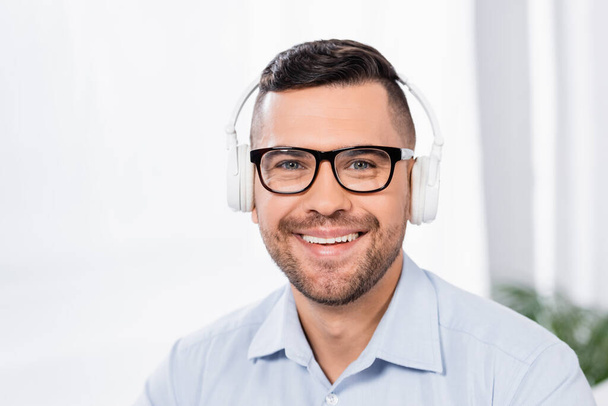 happy businessman in glasses and wireless headphones smiling while looking at camera - Fotografie, Obrázek