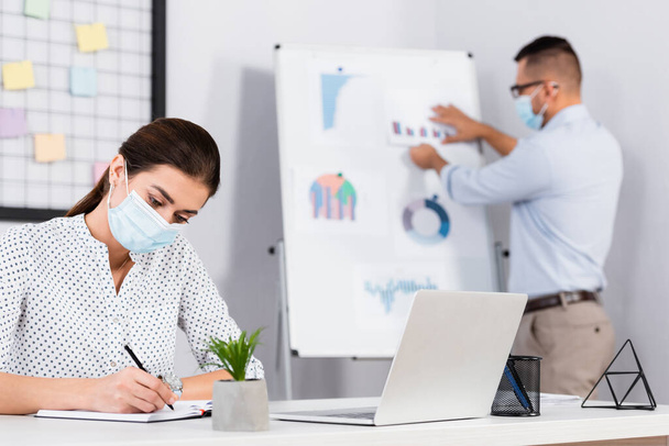 businesswoman in medical mask writing in notebook near businessman on blurred background - Foto, afbeelding