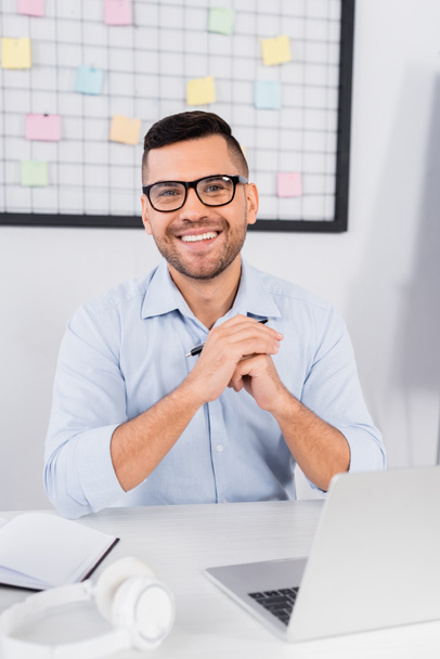alegre hombre de negocios en gafas sonriendo cerca de la computadora portátil en el escritorio - Foto, Imagen
