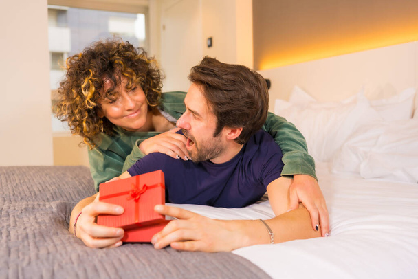 Valentine's Day, a young Caucasian couple in pajamas on the bed, the bride giving a nice surprise with a gift to the boyfriend - Φωτογραφία, εικόνα